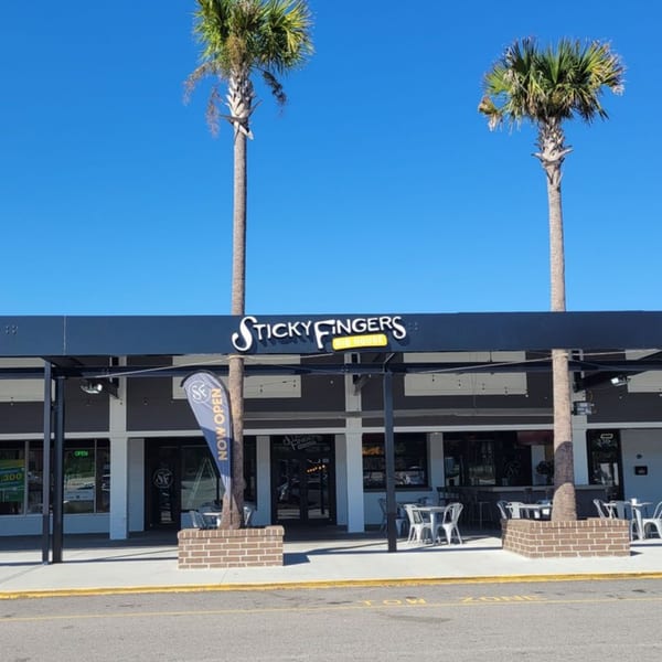 Exterior of Sticky Fingers Rib House with outdoor seating, palm trees, and a "Now Open" banner.