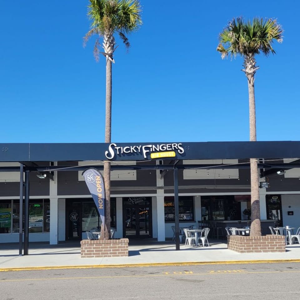 Exterior of Sticky Fingers Rib House with outdoor seating, palm trees, and a "Now Open" banner.