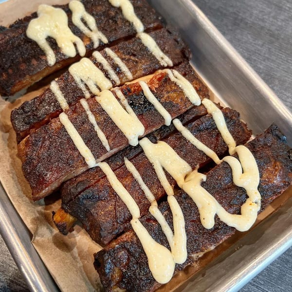 Sticky Fingers Rib House's barbecue ribs, charred and drizzled with a creamy white sauce, in a metal tray.